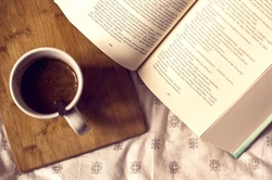 [ai] An open book resting on a light patterned bedspread, with a cup of coffee placed on a wooden board beside it.